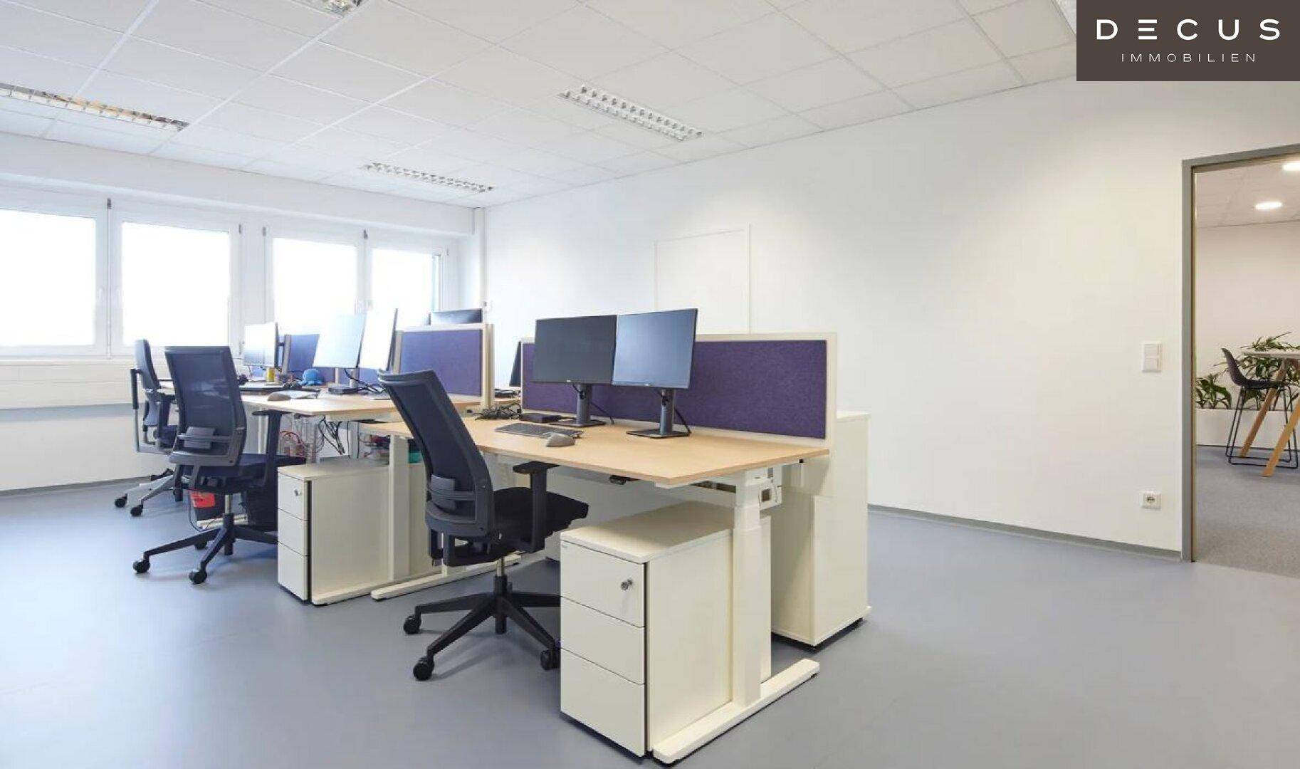 Helles Großraumbüro mit mehreren modernen Arbeitsplätzen, Schreibtischen und großen Fenstern.