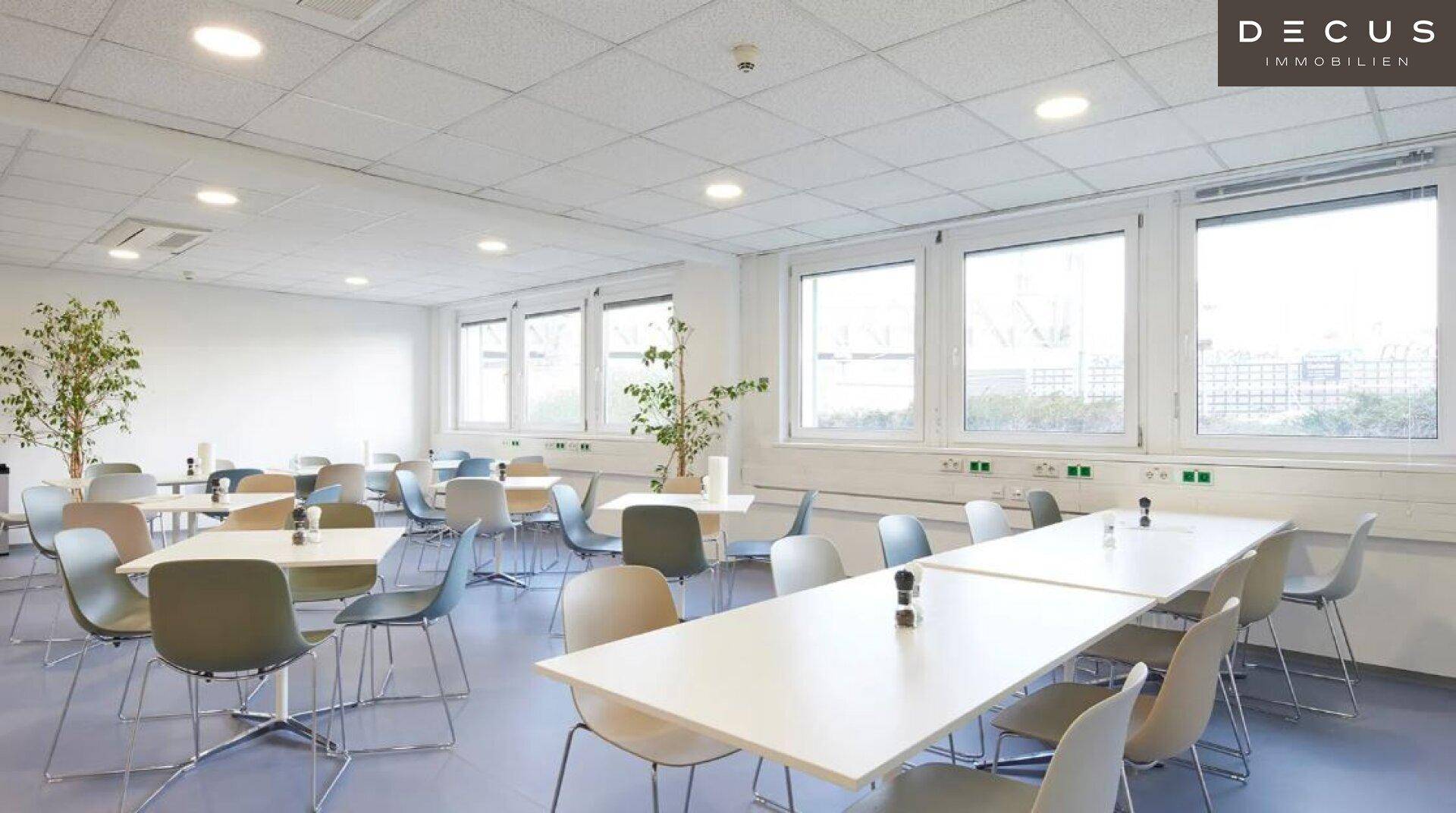 Geräumige Cafeteria oder Pausenraum mit vielen Tischen, Stühlen und großen Fenstern für viel Tageslicht.