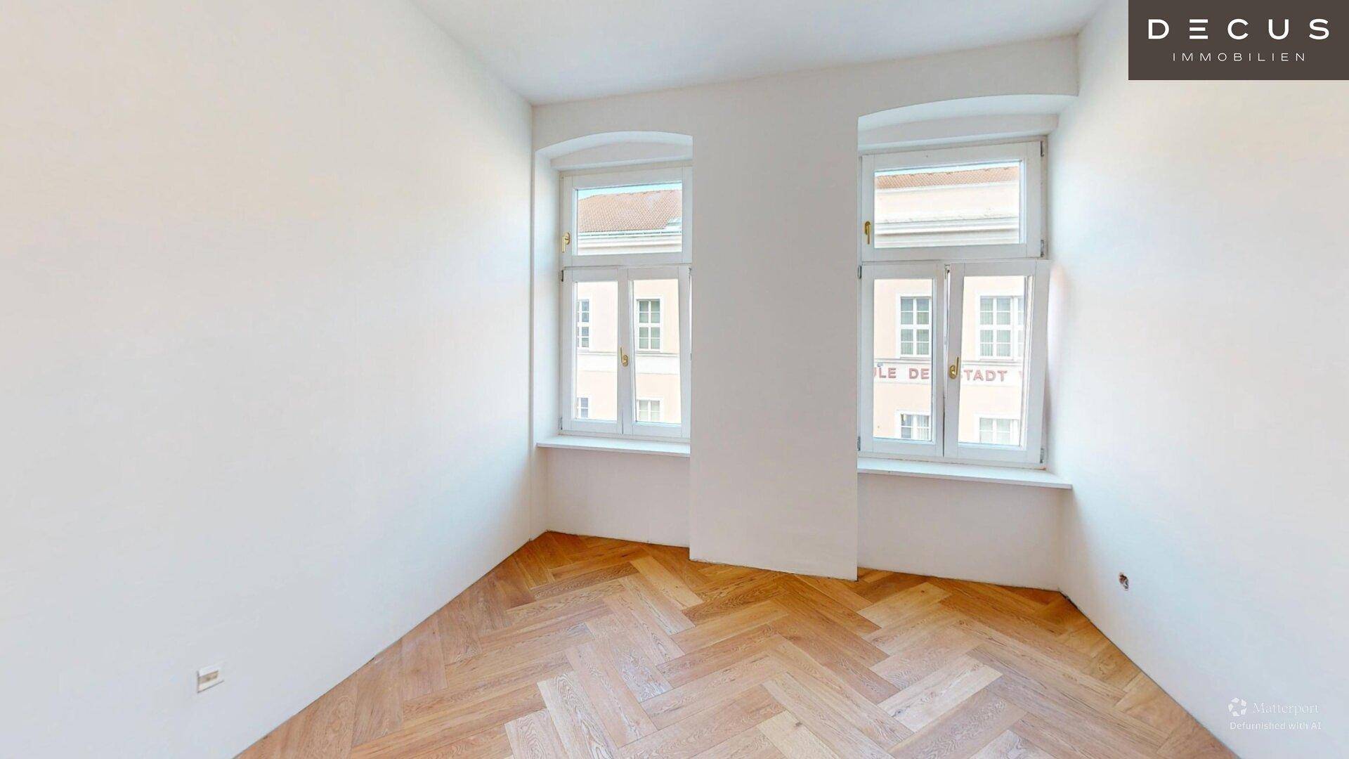Leerer Raum mit Fischgrätparkett und zwei großen Fenstern für viel Tageslicht.