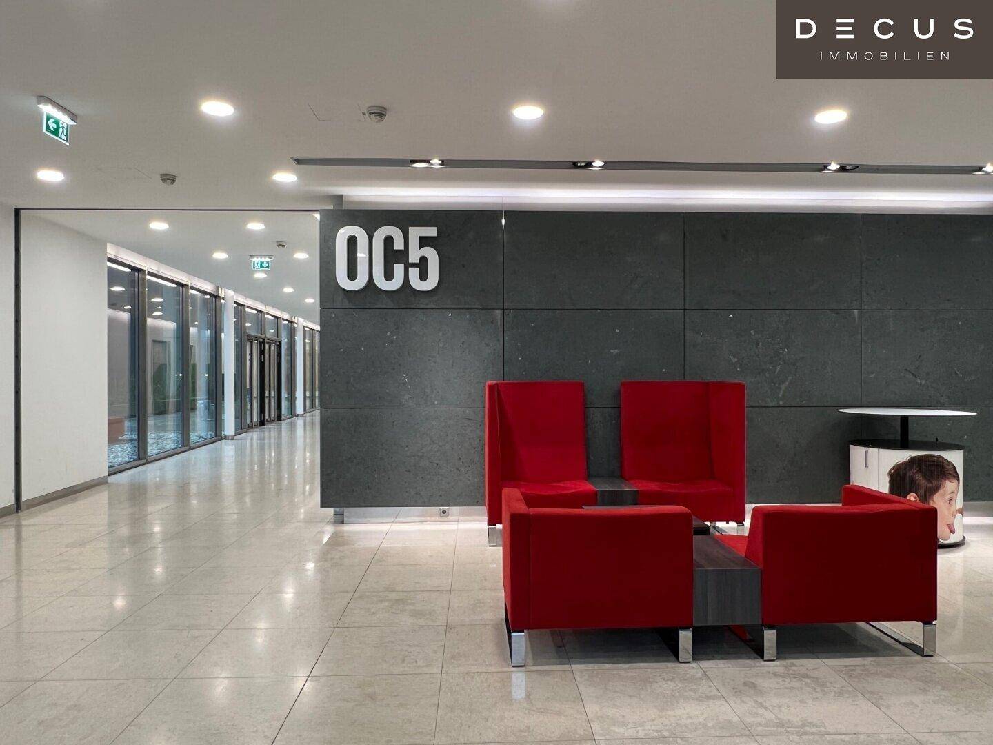 Moderner Loungebereich in einem Bürogebäude mit roten Sesseln und hellem Fliesenboden.