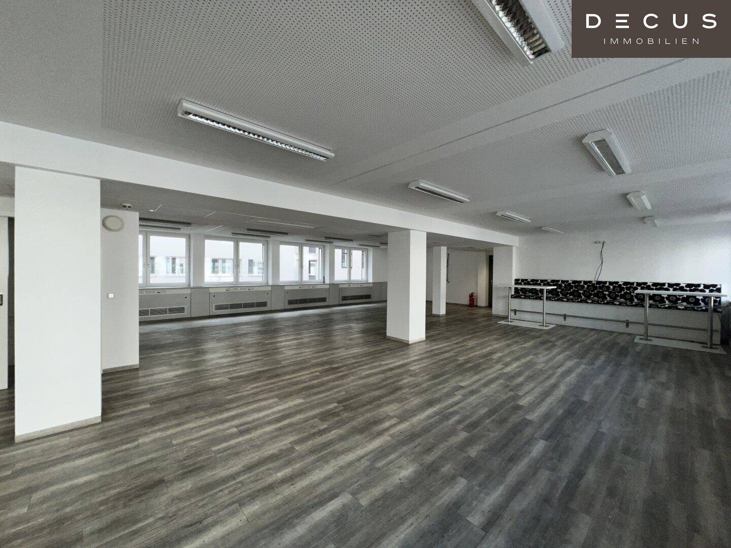 Heller Bürobereich mit durchgehenden Fenstern und grauem Laminatboden, ideal für offene Arbeitsplatzkonzepte.