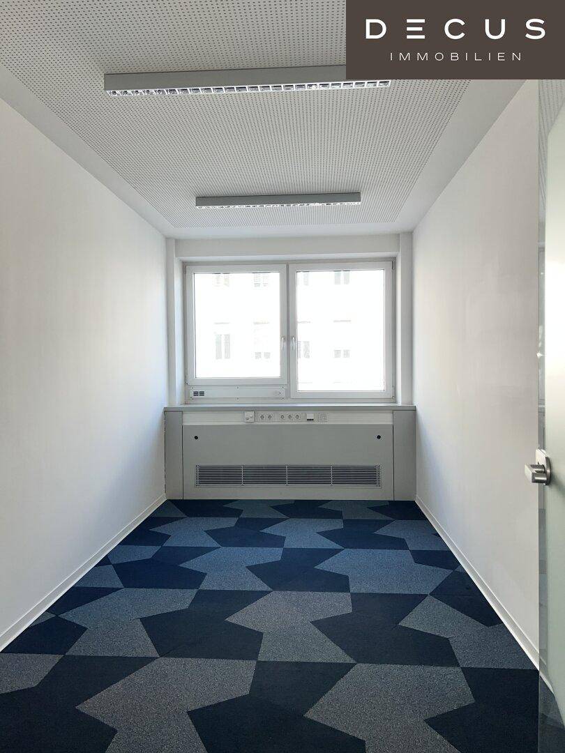 Kleines, helles Büro mit einem Fenster und einem gemusterten Teppichboden, ideal für Einzelarbeit.