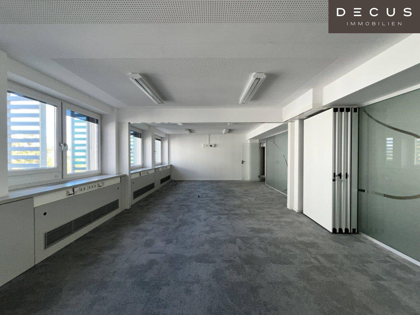 Geräumiges, leeres Büro mit grauem Teppichboden, großen Fenstern und modernen Glasabtrennungen.