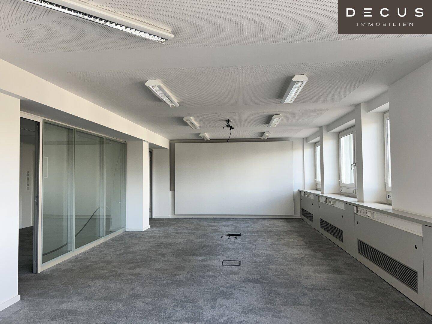 Großer, heller Büroraum mit vielen Fenstern, Teppichboden und einer Projektionsfläche.