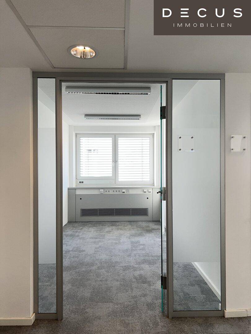 Blick durch eine Glastür in ein helles Büro mit Teppichboden und Jalousien an den Fenstern.