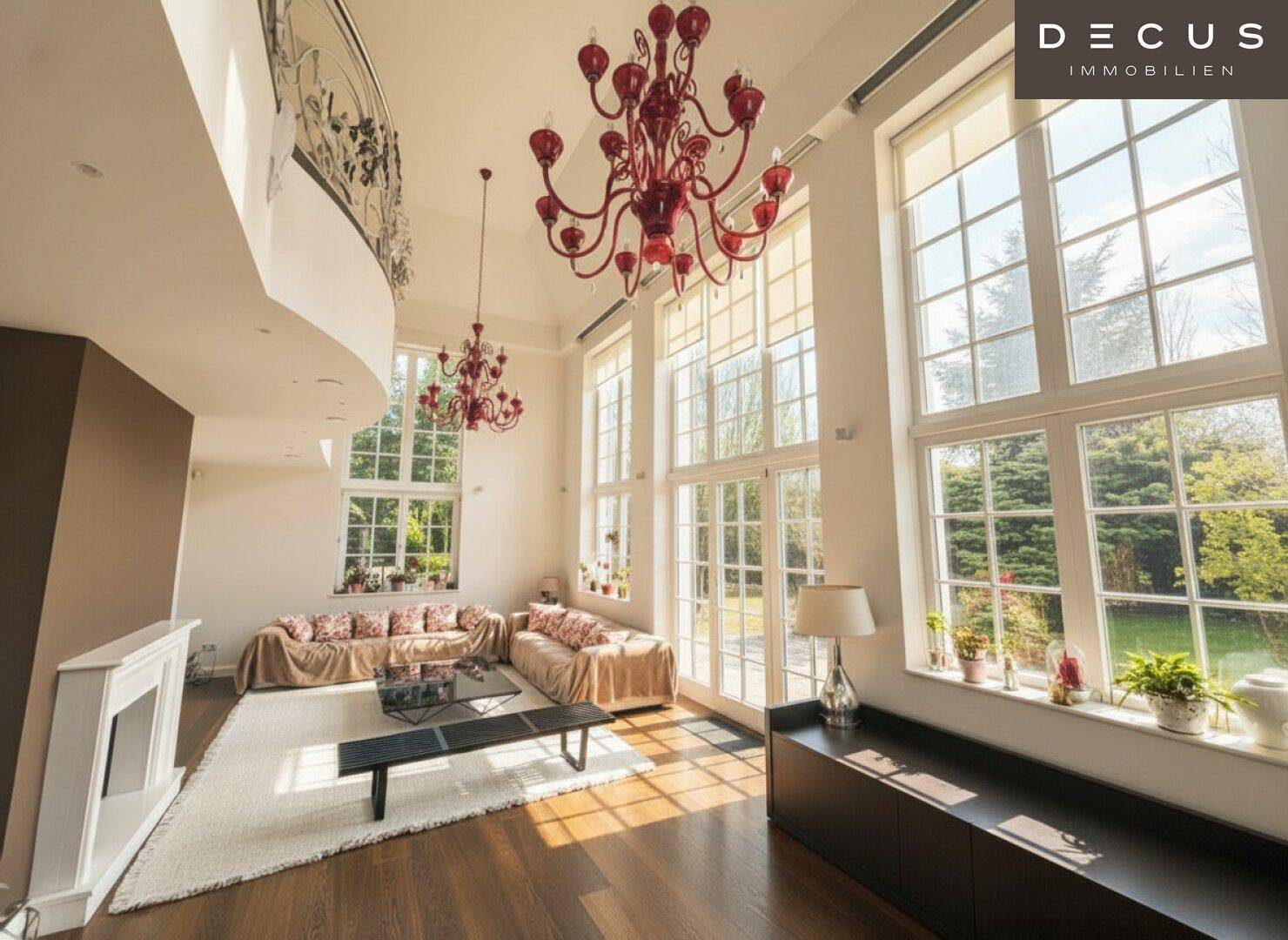 Großzügiges Wohnzimmer mit hohen Decken, bodentiefen Fenstern und zwei roten Kronleuchtern.