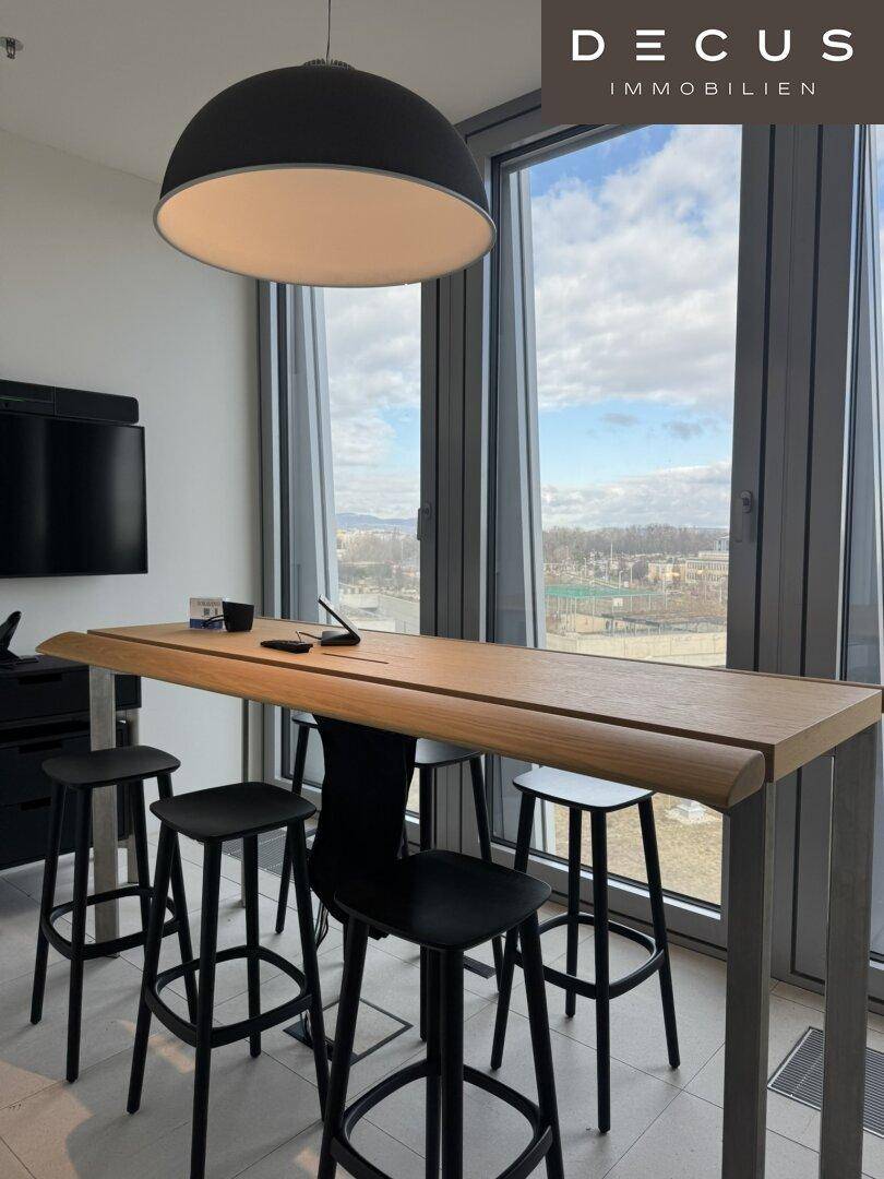Hoher Bartisch mit Hockern vor großen Fenstern, die einen weiten Blick auf die Stadt bieten.