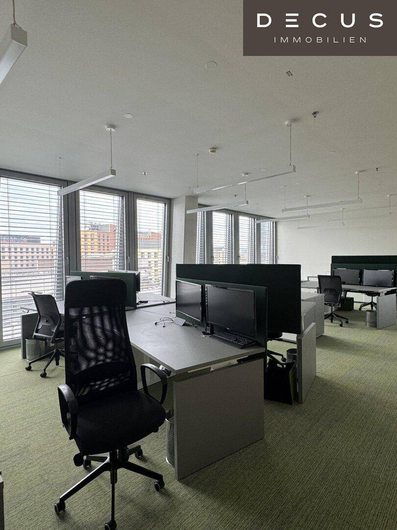 Helles Großraumbüro mit mehreren Arbeitsplätzen, ergonomischen Stühlen und großen Fenstern.
