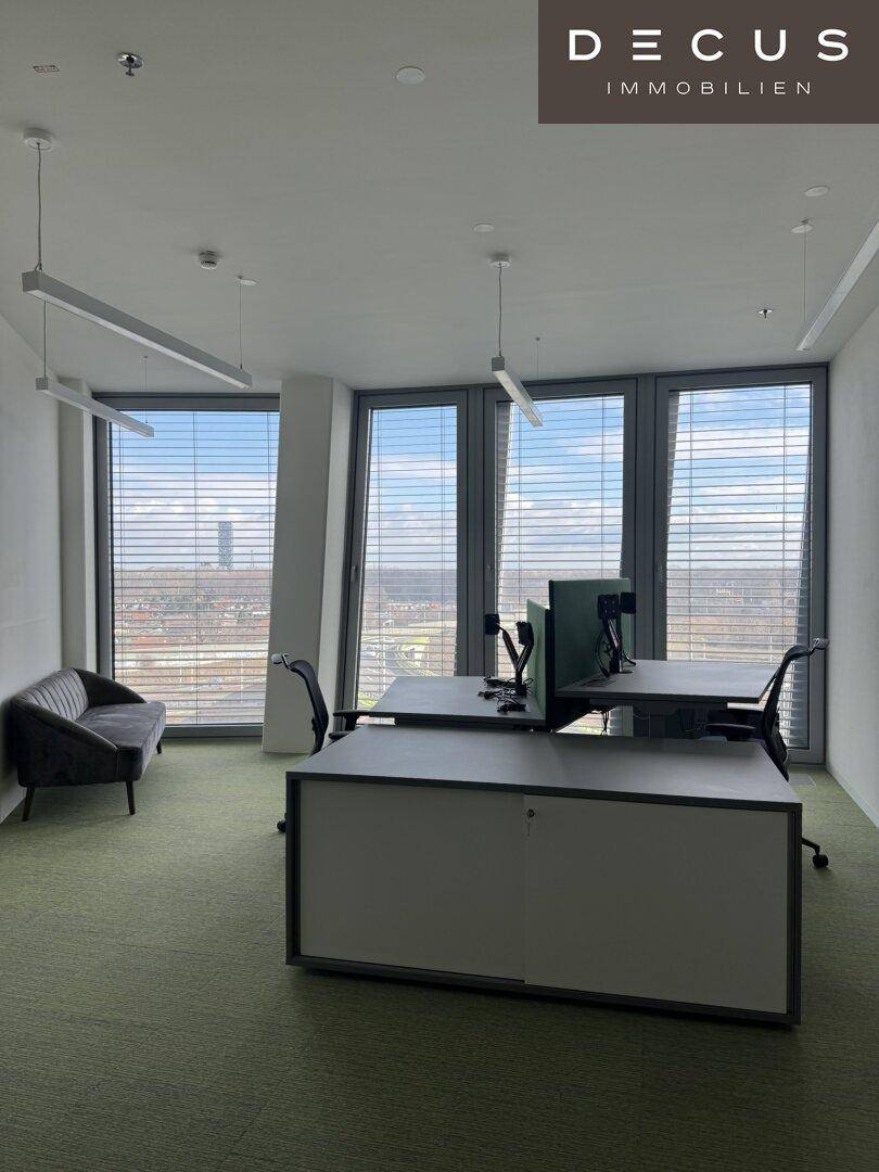 Moderner Bürobereich mit Arbeitsplätzen, einem Sofa und großen Fenstern mit Stadtblick.