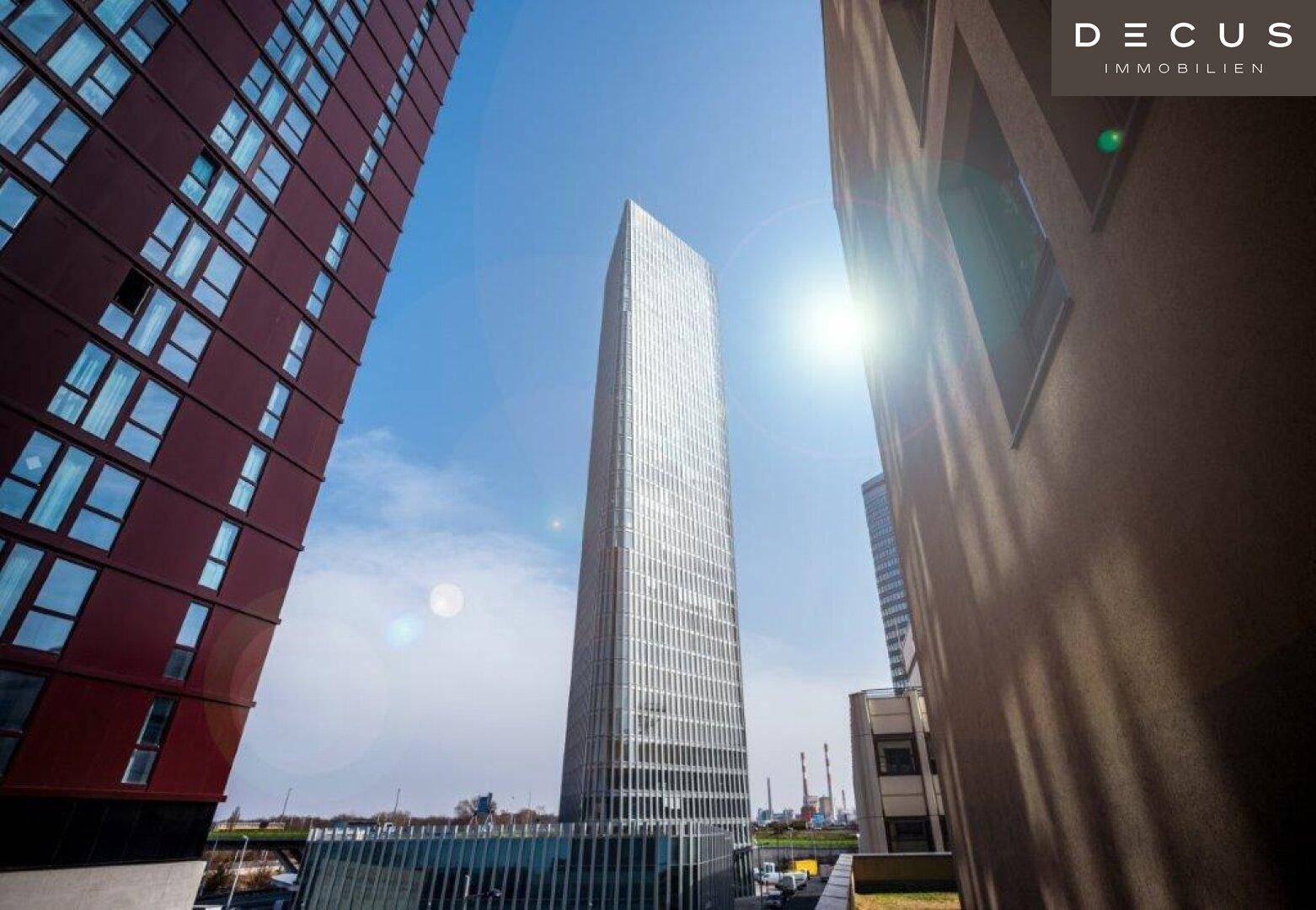 Hohes, modernes Gebäude mit Glasfassade unter blauem Himmel, umgeben von anderen Gebäuden.