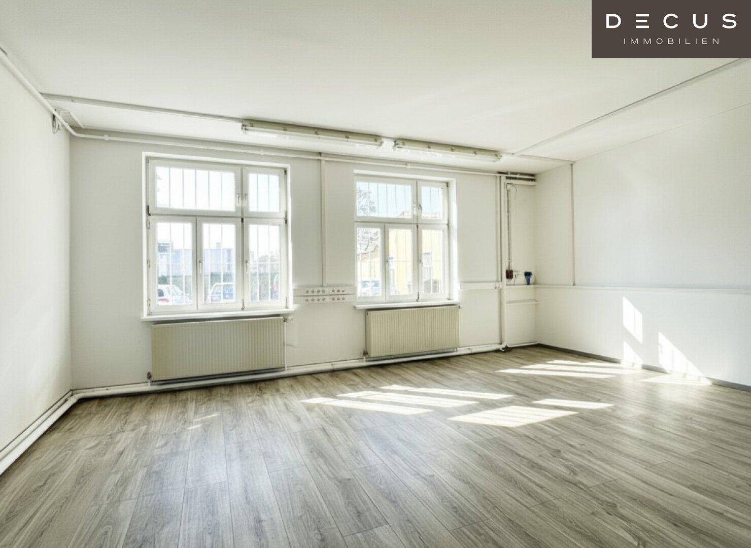 Geräumiger, heller Büroraum mit Laminatboden, weißen Wänden und zwei großen Fenstern.