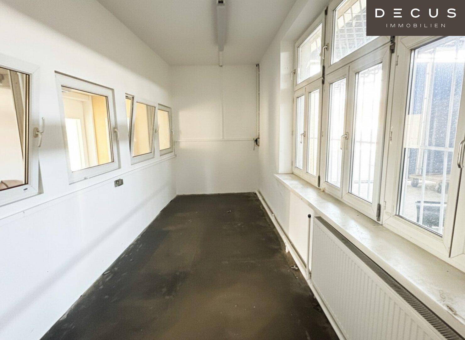 Schmaler Raum mit dunklem Boden, weißen Wänden und mehreren Fenstern, teilweise vergittert.