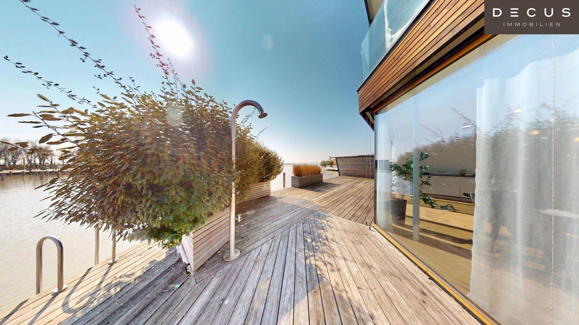 Sonnige Holzterrasse direkt am Wasser mit Außendusche und üppiger Bepflanzung.