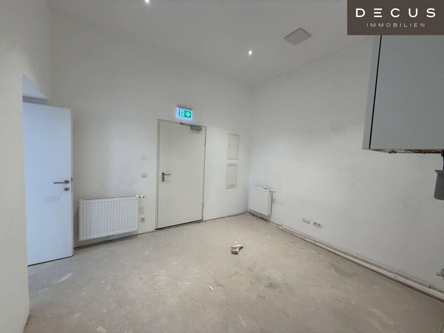 Leerer Gewerberaum mit weißen Wänden, Heizkörpern und einer Tür, die zu weiteren Räumen führt.