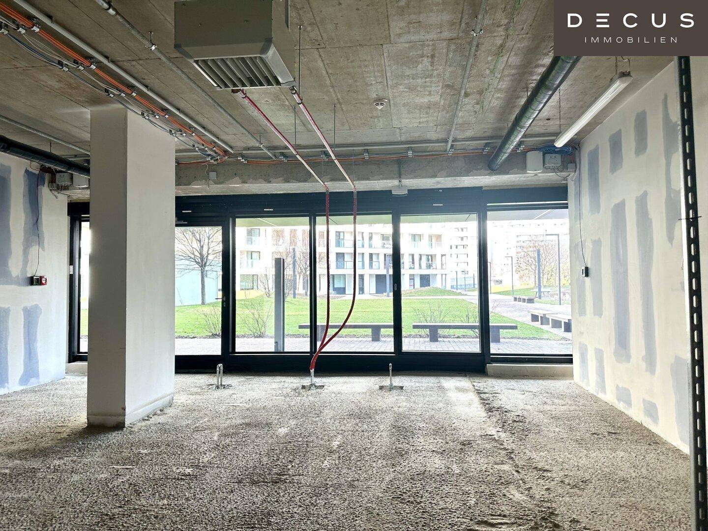 Unfertiger Gewerberaum mit freiliegenden Deckeninstallationen und großen Fenstern mit Blick ins Grüne.