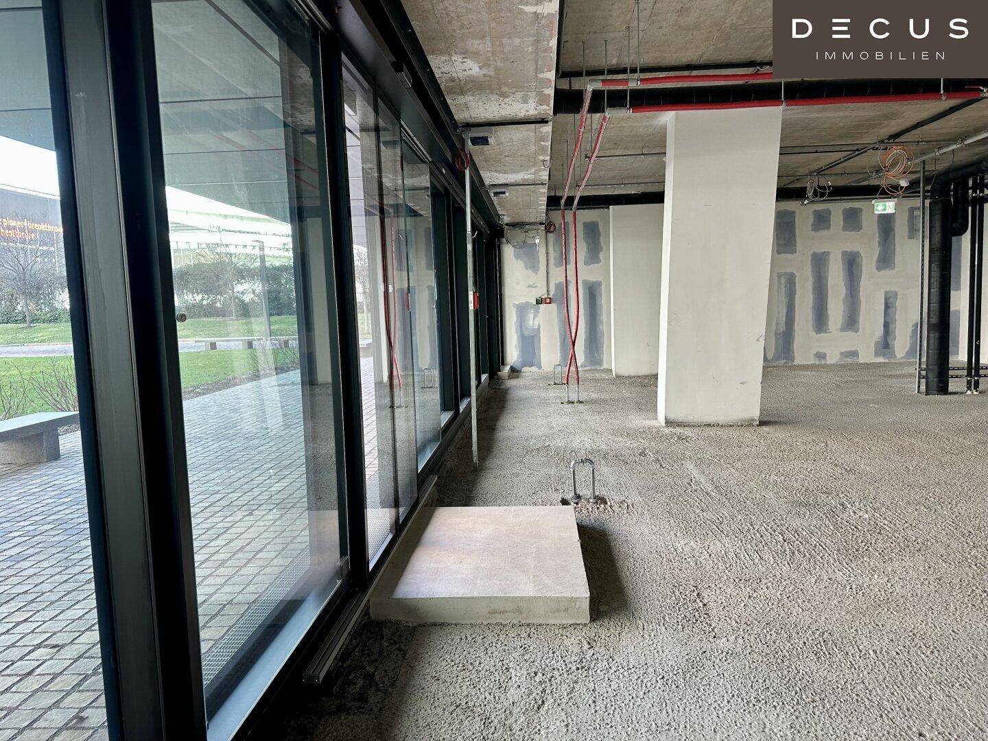 Unfertiger Innenraum eines Bürogebäudes mit großen Fenstern und Blick nach draußen auf den Gehweg.