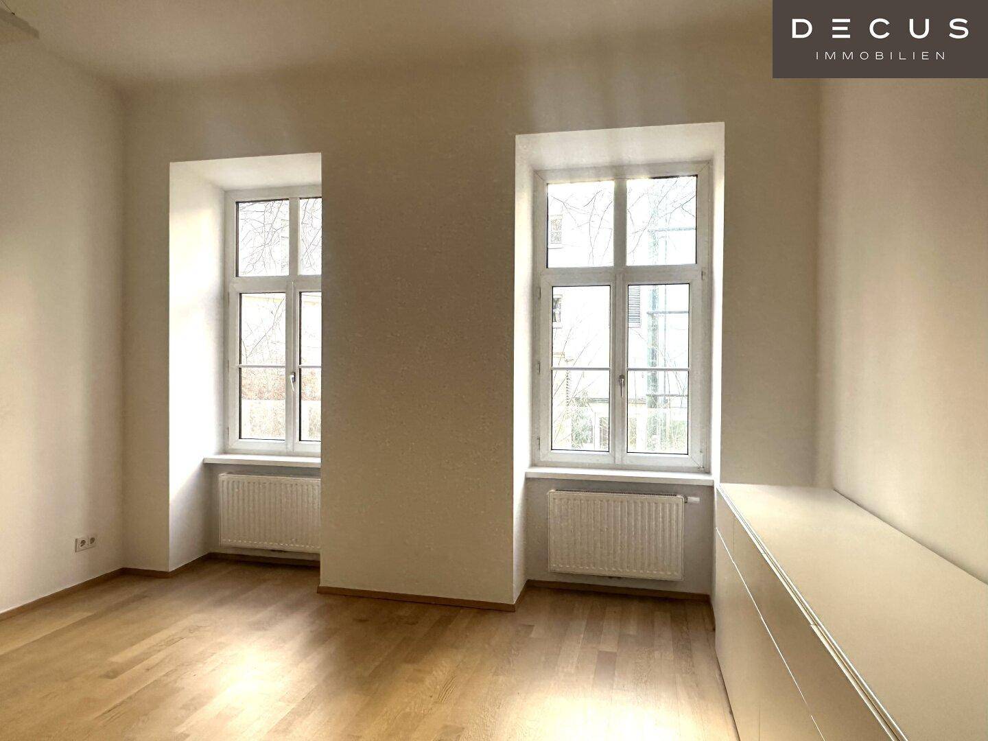 Heller Büroraum mit zwei großen Fenstern, Holzfußboden und einem langen weißen Sideboard.