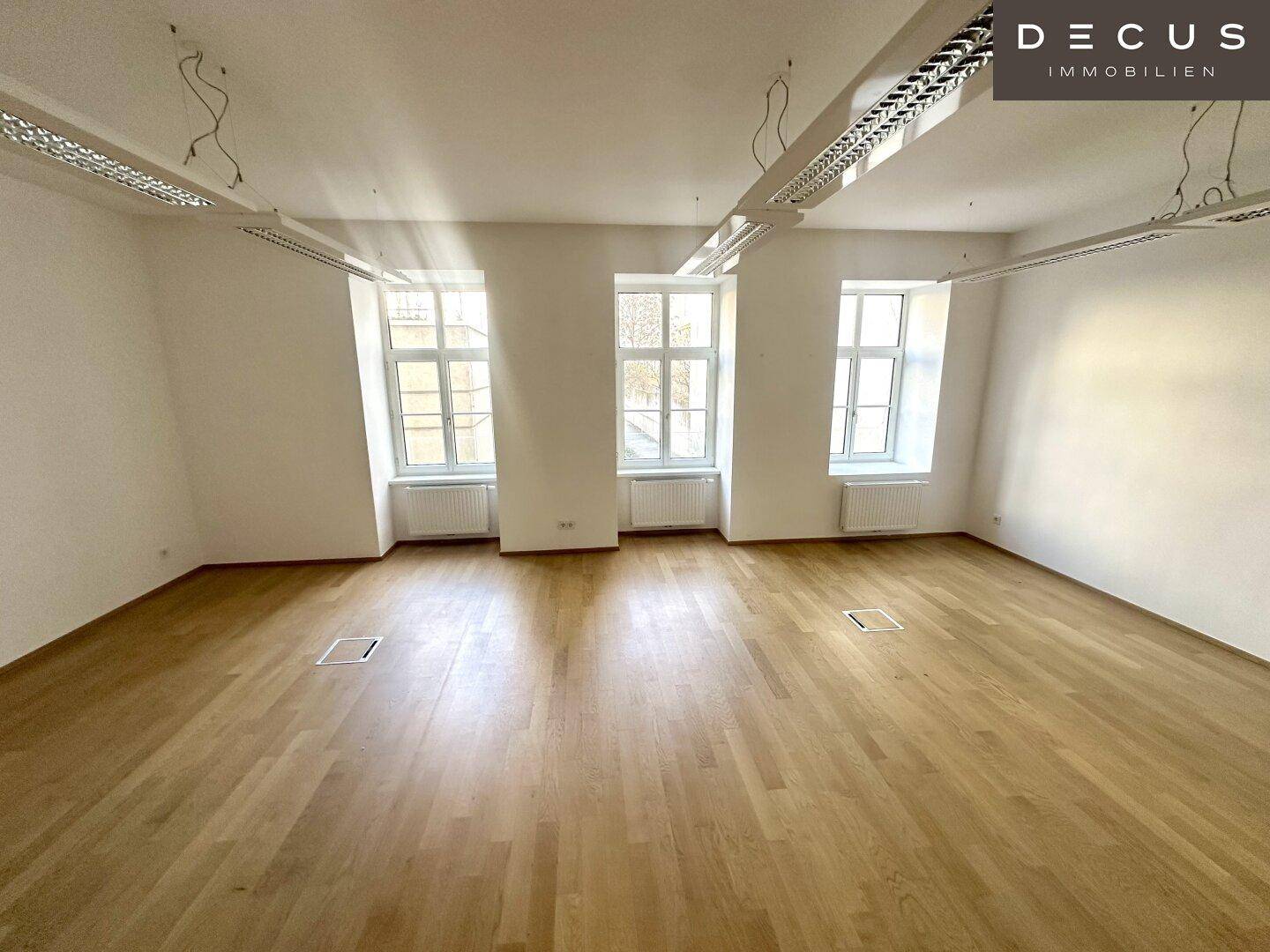Großer, heller Büroraum mit drei Fenstern, Parkettboden und Deckenleuchten.