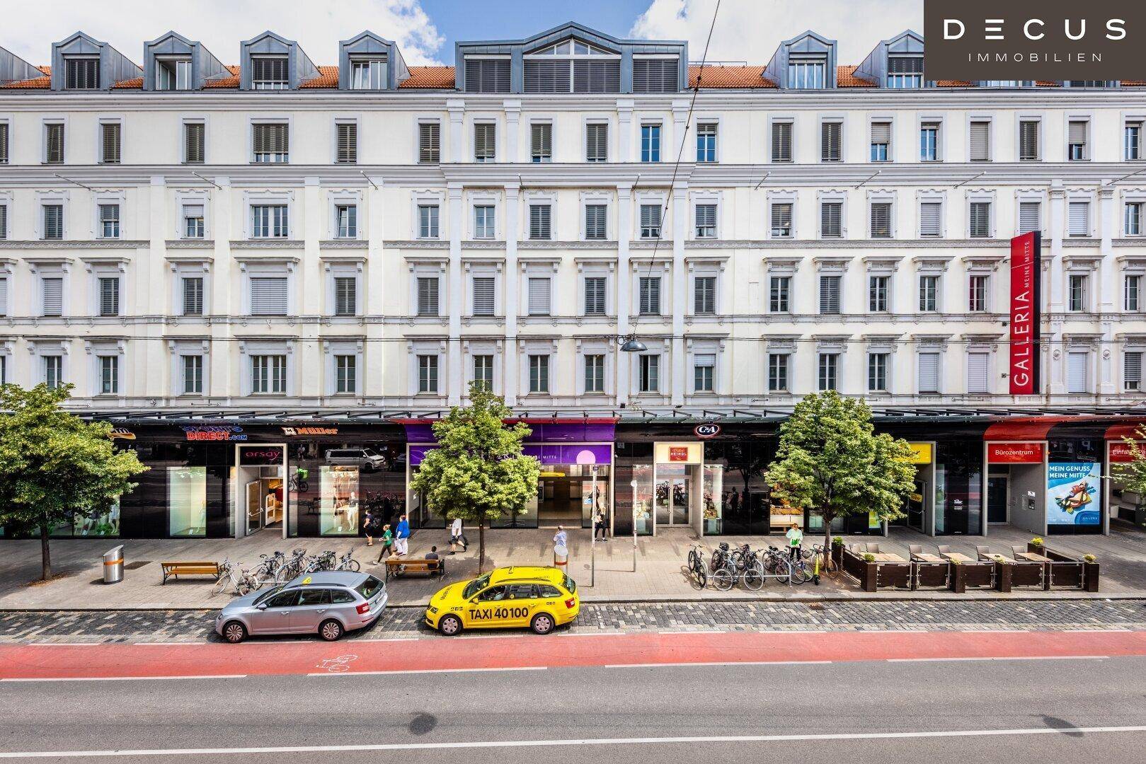 Ansicht einer belebten Stadtfassade mit Geschäften im Erdgeschoss und Straßenverkehr davor.