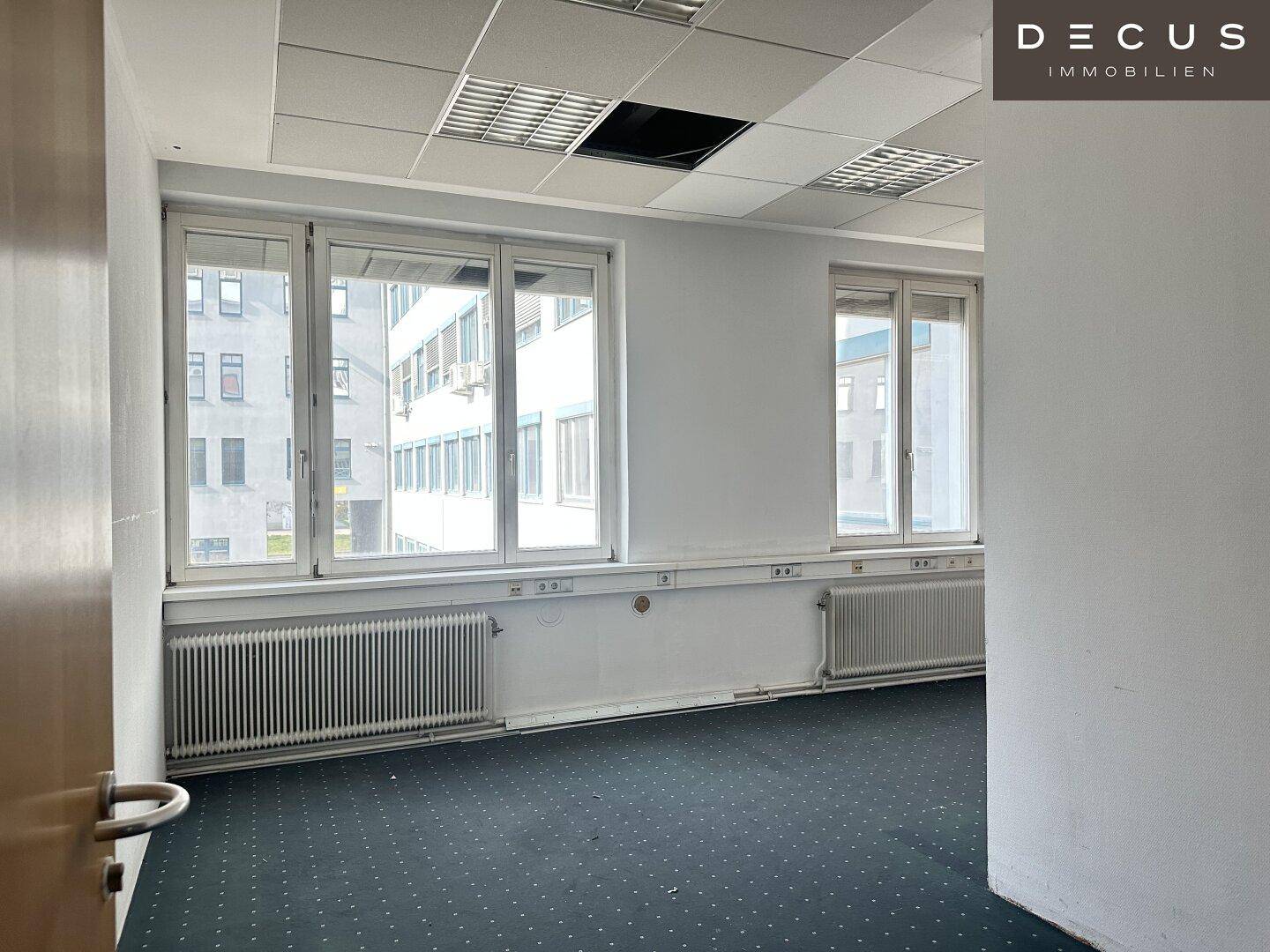 Helles Büro mit Teppichboden, großen Fenstern und einer Rasterdecke, bietet viel Tageslicht.
