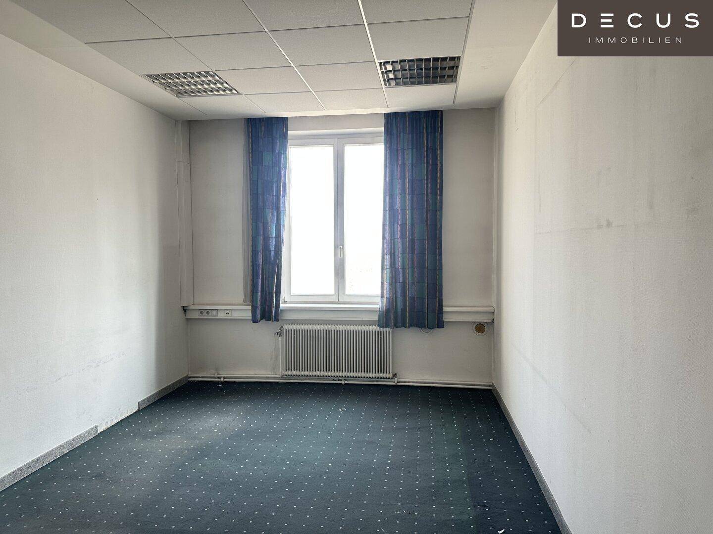 Kleineres Büro mit dunklem Teppichboden, Fenster mit Vorhängen und Heizkörper, ideal für Einzelarbeit.