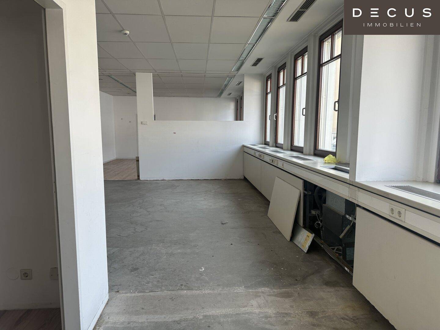 Heller Büroraum mit Trennwänden, Fensterfront und vorbereiteten Installationen unter den Fenstern.
