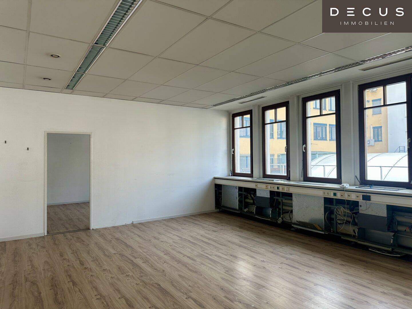 Leerer Büroraum mit Laminatboden, großen Fenstern und einer offenen Tür zu einem weiteren Bereich.