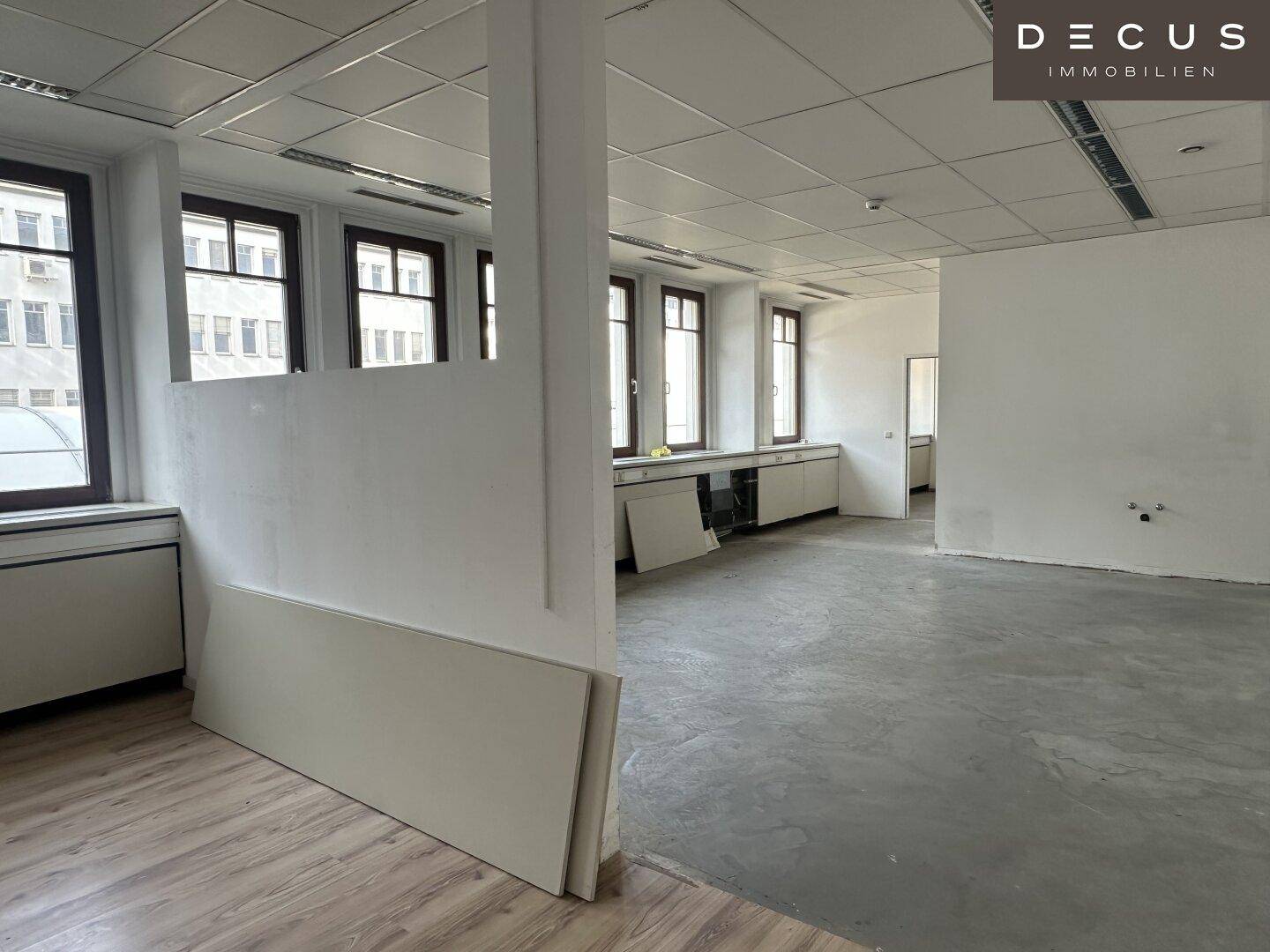 Offener Bürobereich mit Trennwand, großen Fenstern und einer Kombination aus Laminat- und Betonboden.