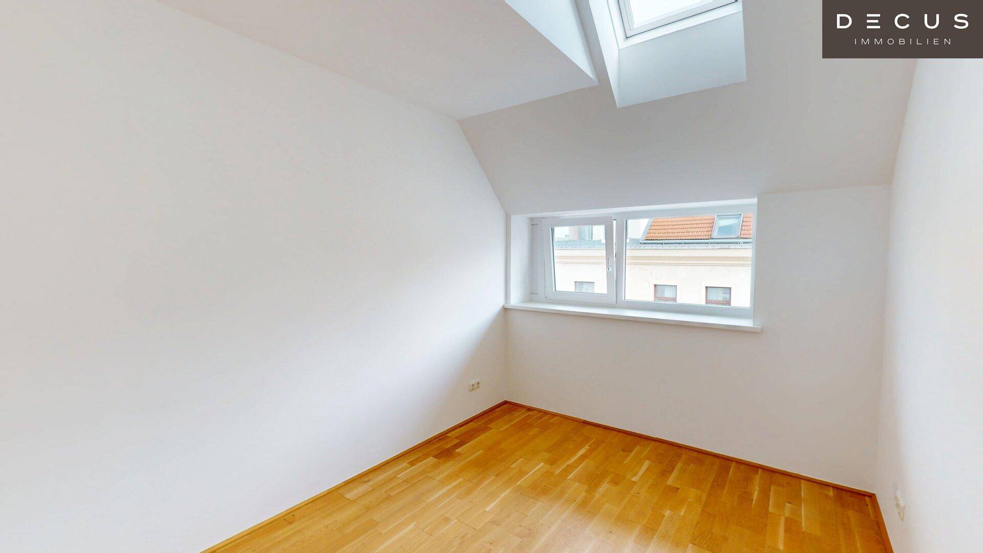 Geräumiges Zimmer im Dachgeschoss mit Holzboden und einem Fenster mit Blick auf die Dächer.