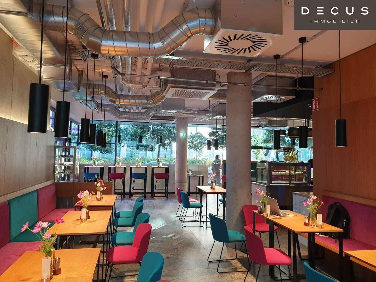 Modernes Café mit farbenfrohen Sitzgelegenheiten, sichtbaren Lüftungsrohren und viel Tageslicht.