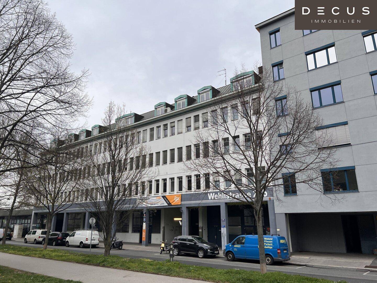 Weitwinkelansicht der modernen Gebäudefassade entlang der Straße mit Bäumen und Fahrzeugen.