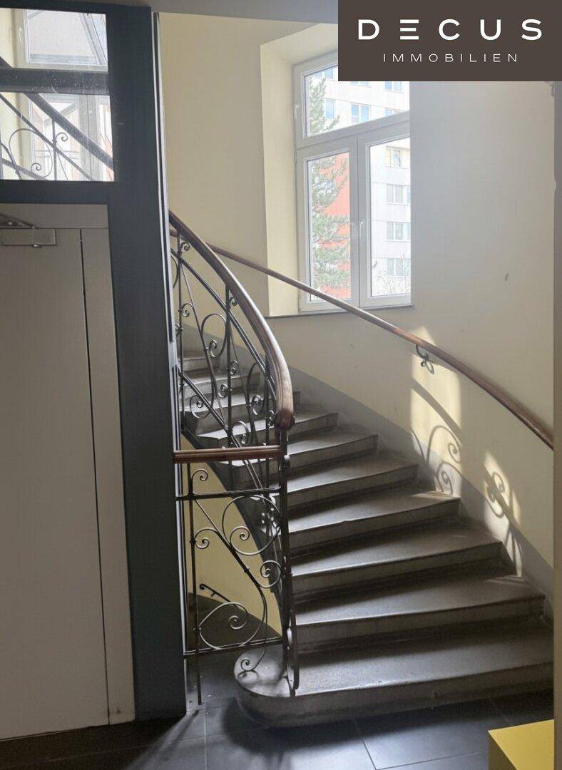 Historisches Treppenhaus mit schmiedeeisernem Geländer und großen Fenstern, das viel Tageslicht hereinlässt.