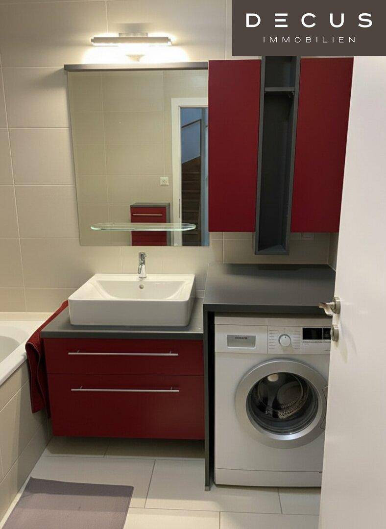 Modernes Badezimmer mit Waschbecken, Spiegel und integrierter Waschmaschine, in hellen Farbtönen gehalten.