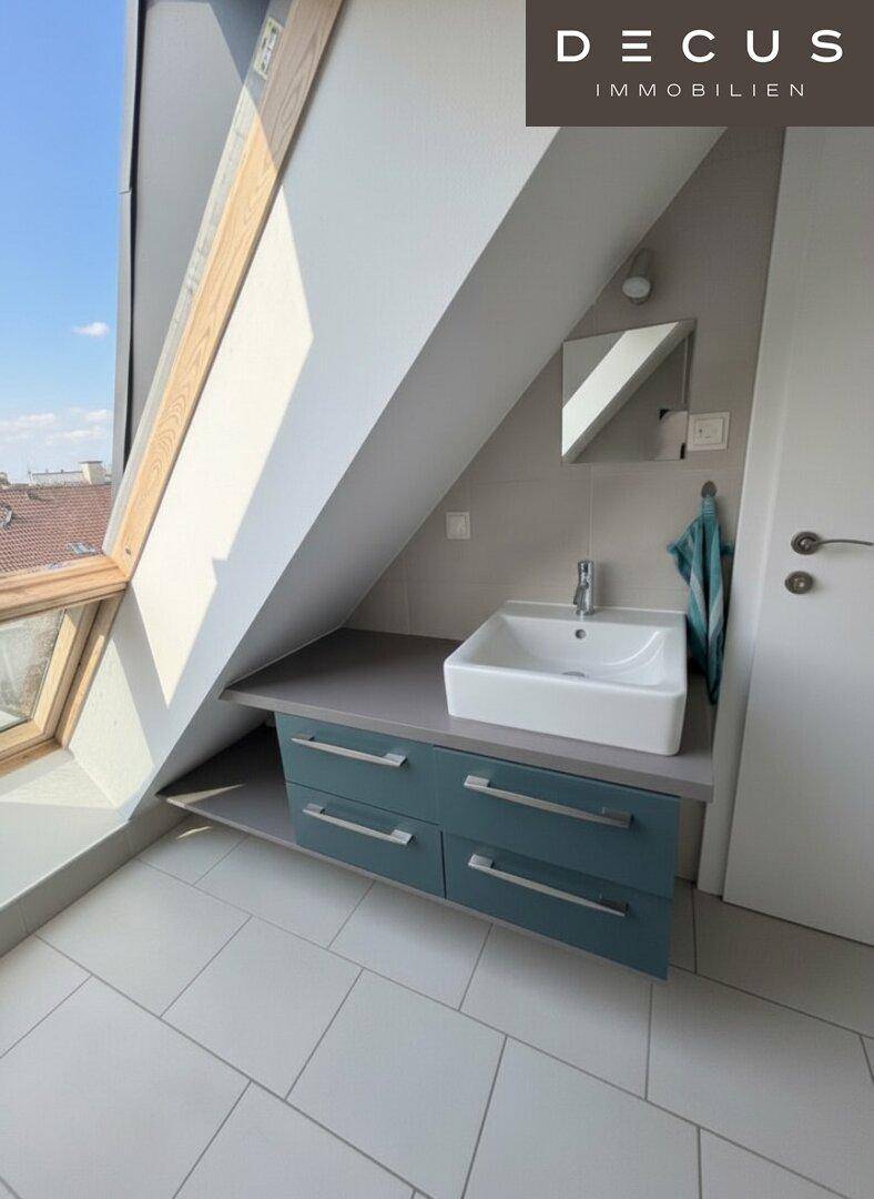 Helles Badezimmer mit Waschbecken, Dachfenster und modernen blauen Unterschränken unter der Dachschräge.