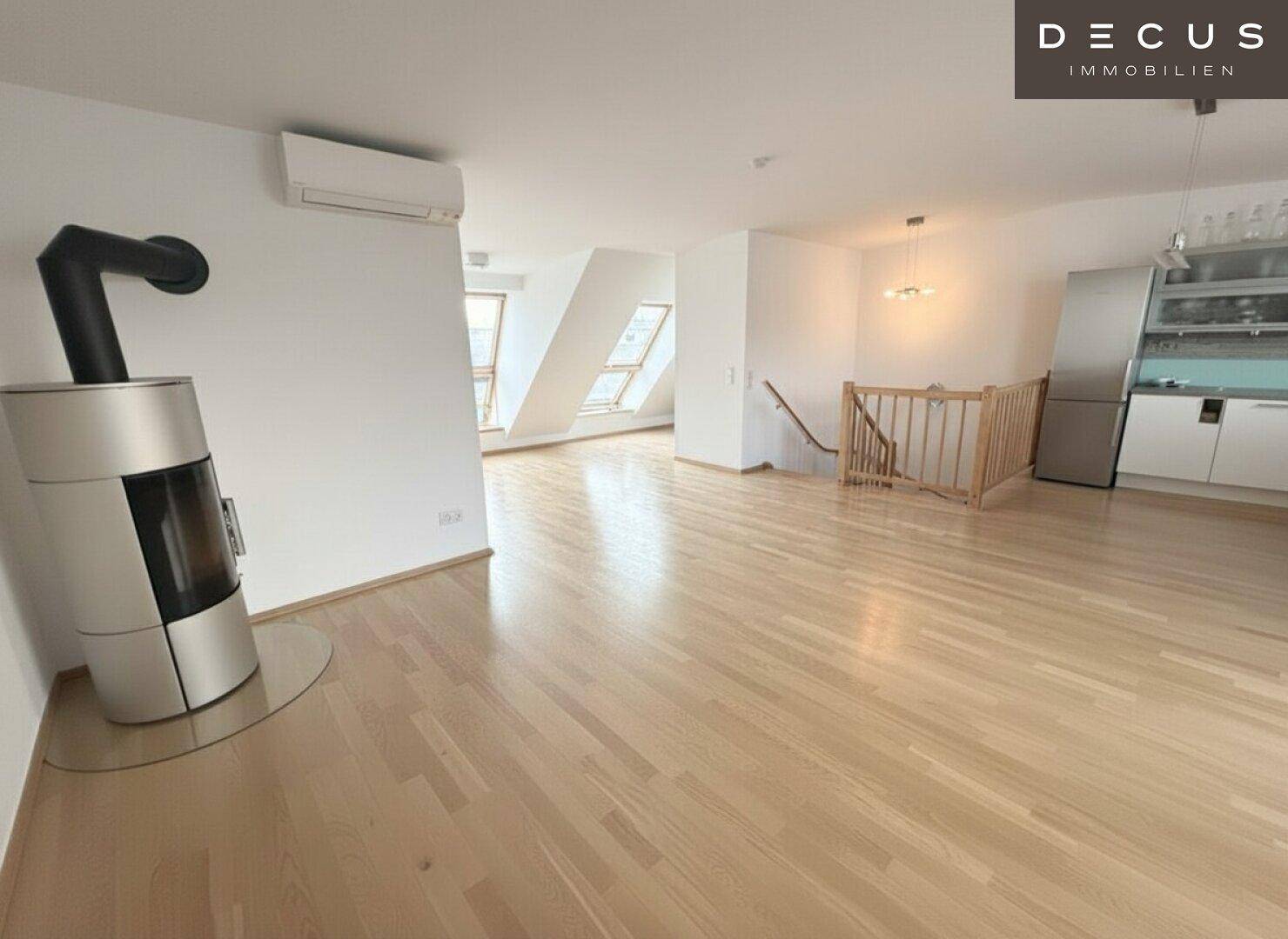 Heller offener Wohnbereich mit Holzboden, Kaminofen und Blick auf die Küche, unter Dachschrägen.