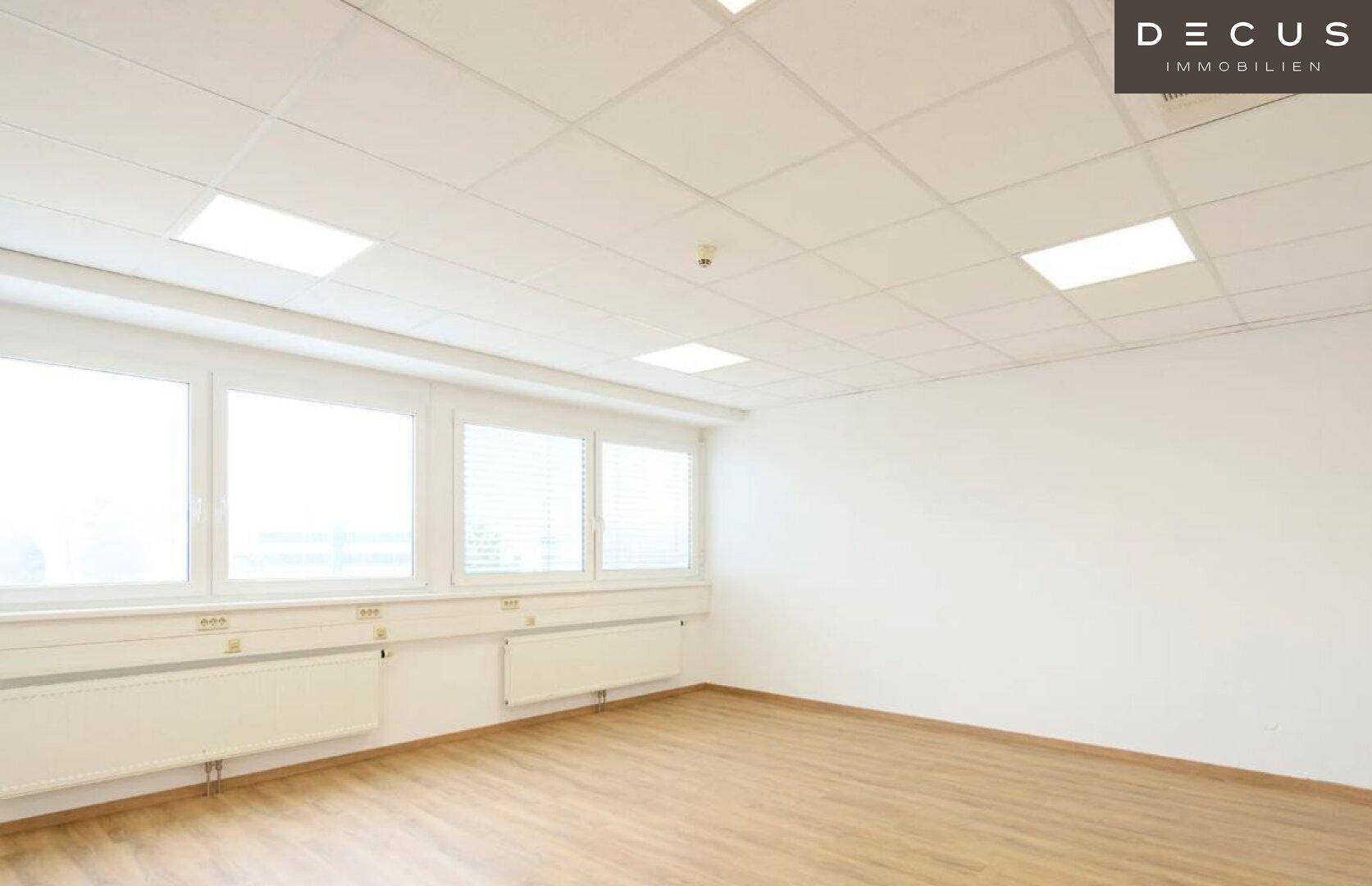 Leerer Büroraum mit hellem Holzboden, großen Fenstern und Deckenbeleuchtung.