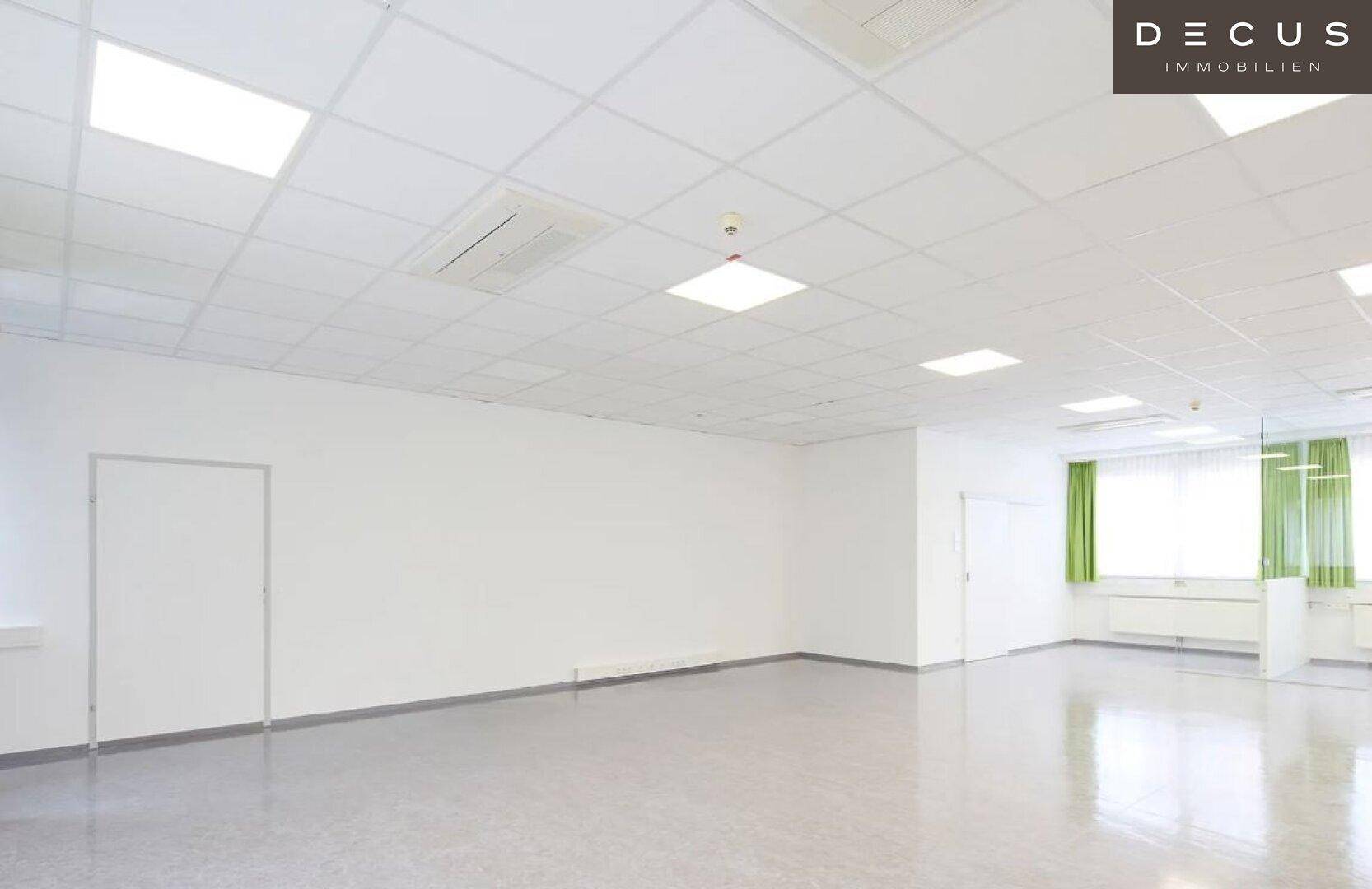 Geräumiger Büroraum mit grauem Vinylboden, weißen Wänden und Deckenplatten mit integrierter Beleuchtung.