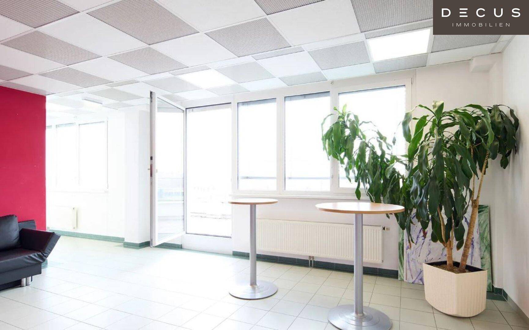 Heller Loungebereich mit Stehtischen, Pflanzen und großen Fenstern, die viel Tageslicht hereinlassen.
