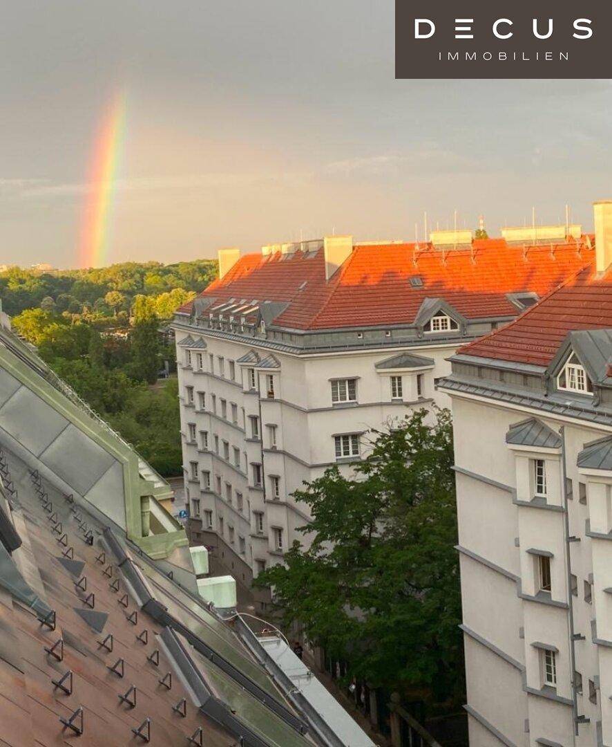 Panoramablick über die Stadt mit traditionellen Dächern, Bäumen und einem Regenbogen am Horizont.