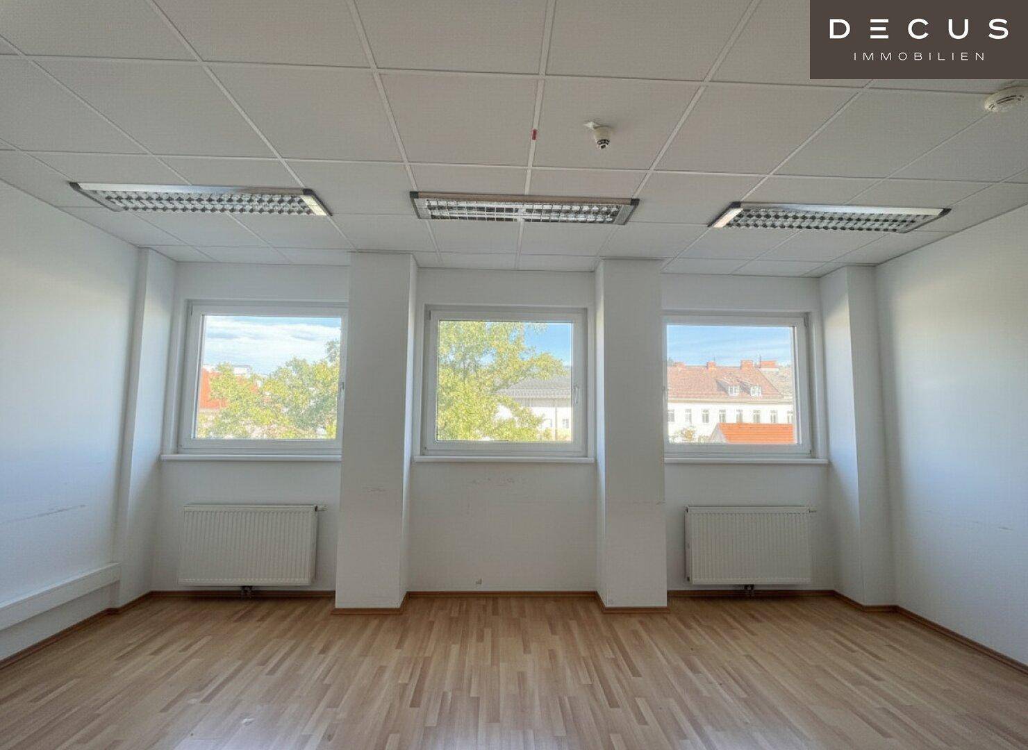 Heller Büroraum mit drei großen Fenstern und Holzfußboden, bietet eine angenehme Arbeitsatmosphäre.