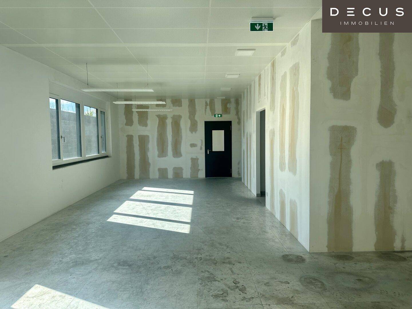 Geräumiger Büroraum im Rohbau mit Betonboden und großen Fenstern, die viel Licht hereinlassen.