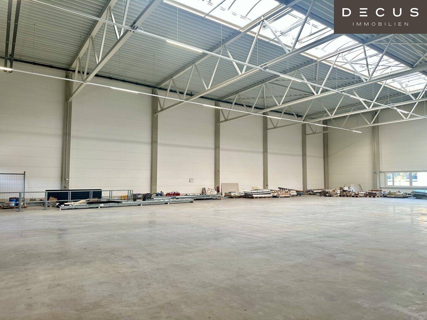 Weitläufige, leere Lagerhalle mit hohen Decken, Metallkonstruktion und viel Tageslicht.