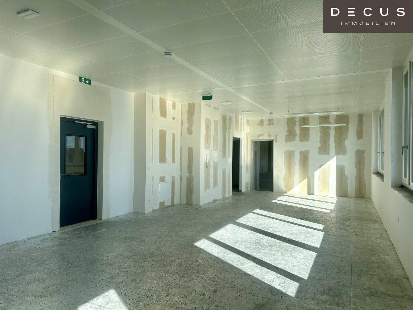 Unfertiger Büroraum mit Betonboden, abgehängter Decke und teilweise verputzten Wänden.