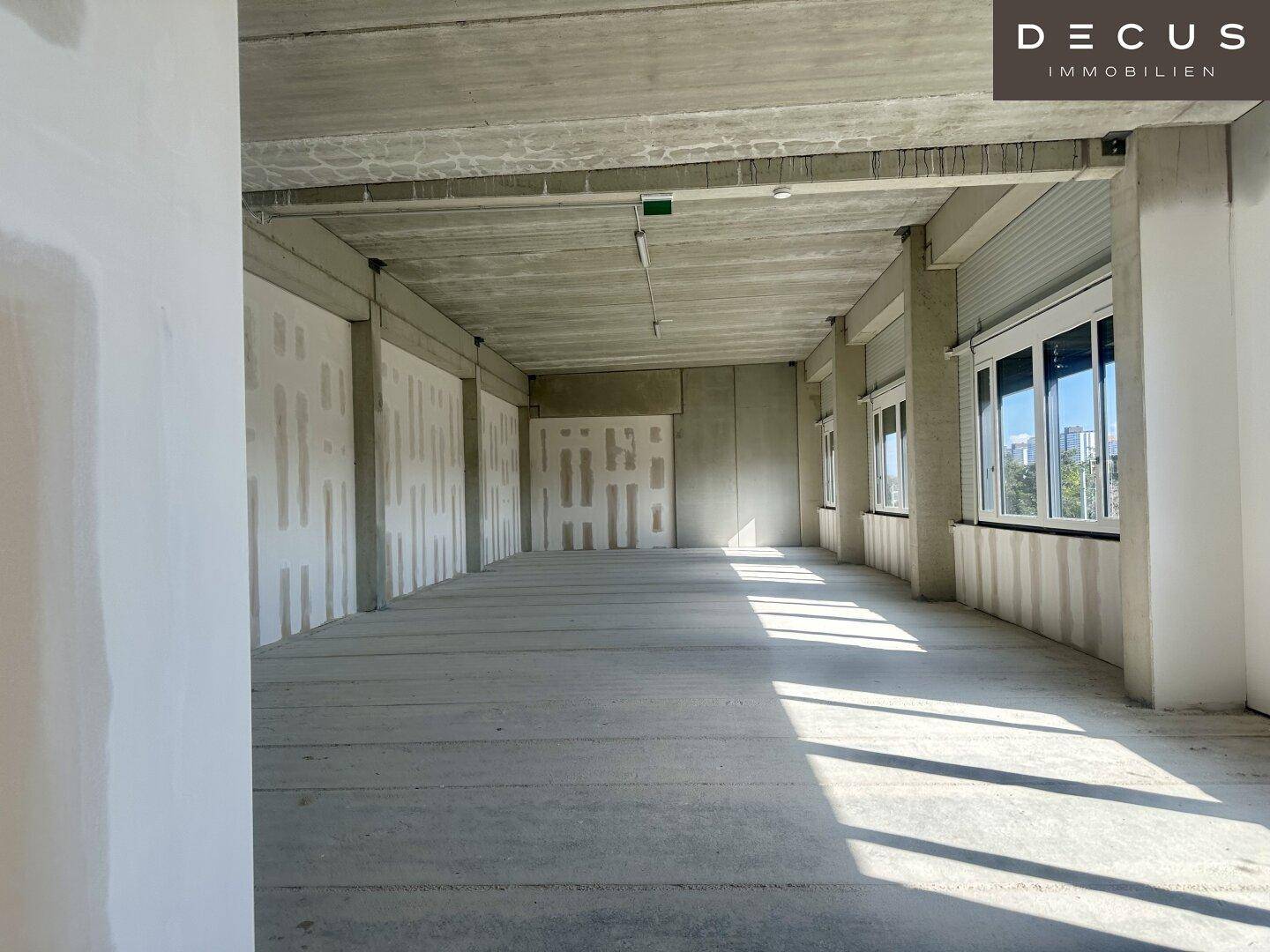 Büroraum im Rohbau mit sichtbarer Betondecke, Säulen und Fenstern mit Rollläden.