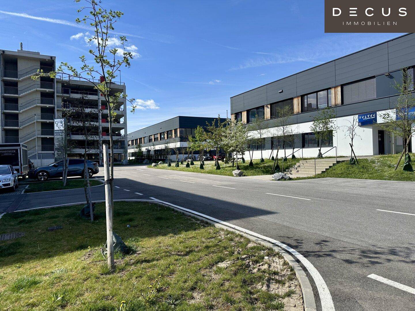 Ansicht einer modernen Gewerbezone mit mehreren Gebäuden, Parkplätzen und begrünten Flächen unter blauem Himmel.