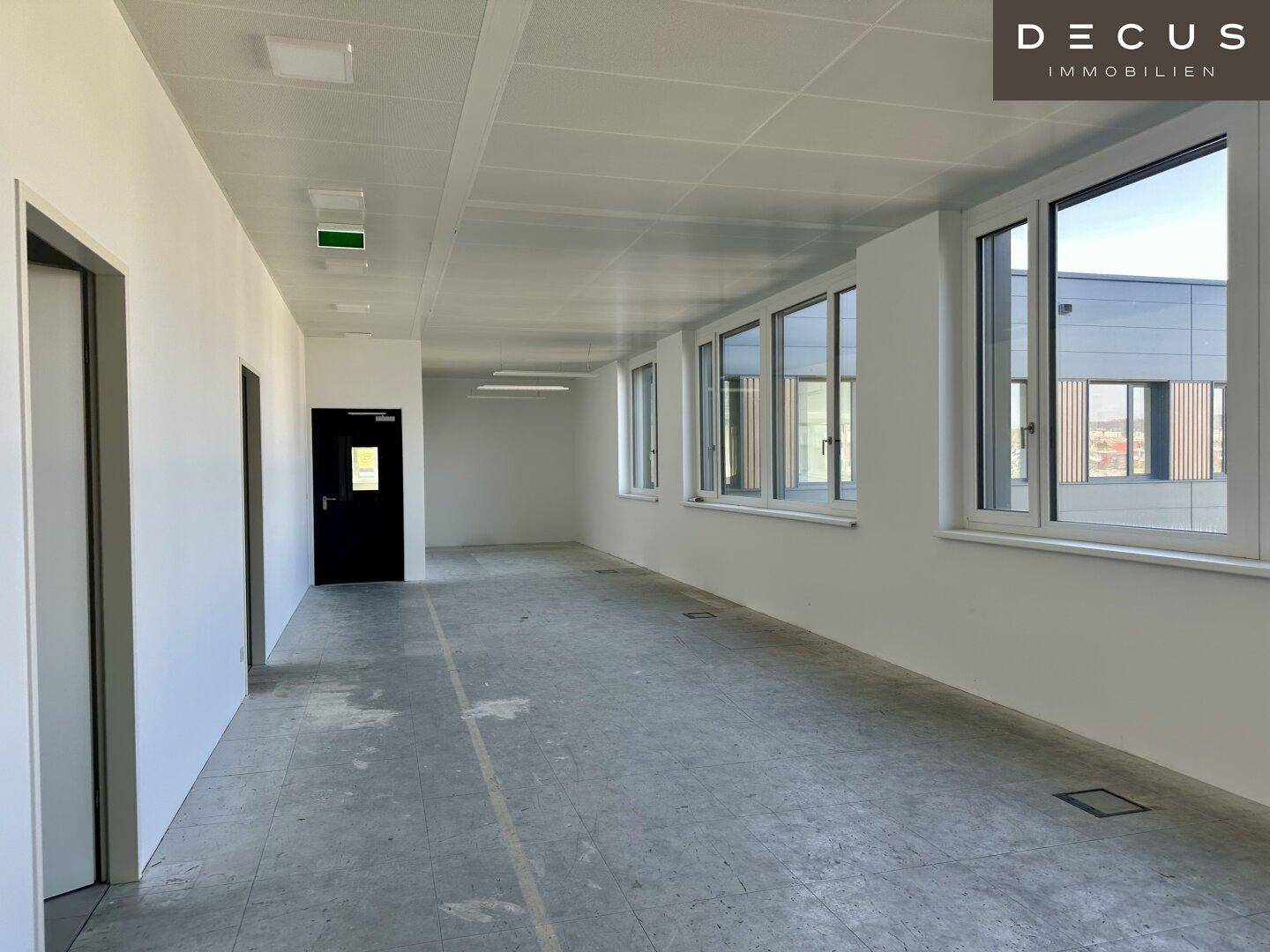 Langer, offener Bürobereich mit vielen Fenstern entlang der Wand, die für eine helle Arbeitsumgebung sorgen.