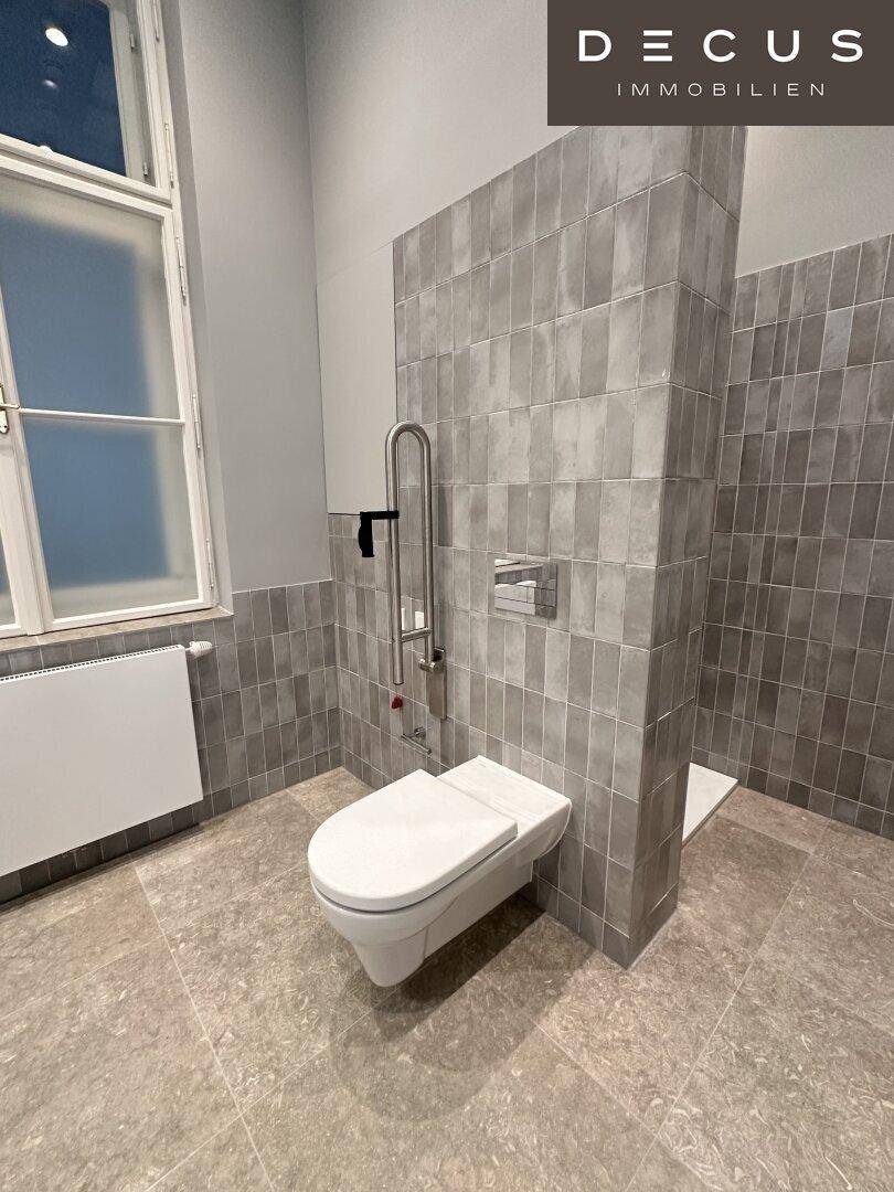 Modernes Badezimmer mit grauen Fliesen, wandhängender Toilette und Haltegriffen für verbesserte Barrierefreiheit.