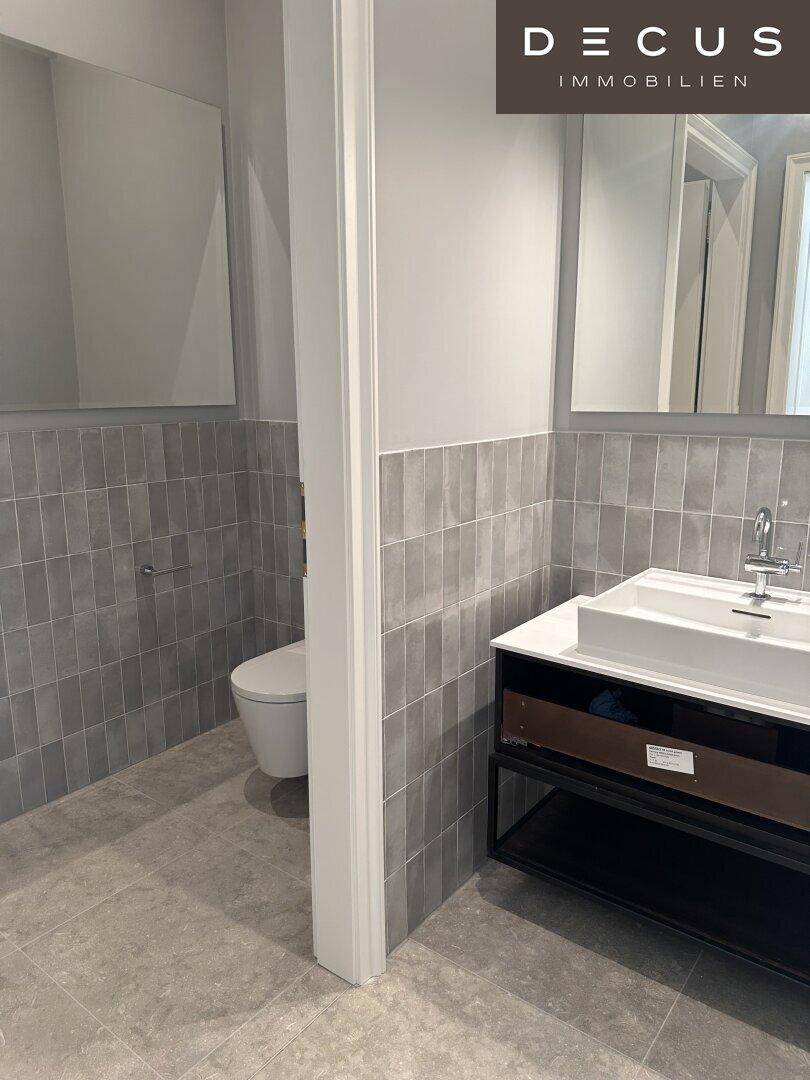 Modernes Badezimmer mit stilvollem Waschtisch, großem Spiegel und eleganten grauen Wandfliesen.