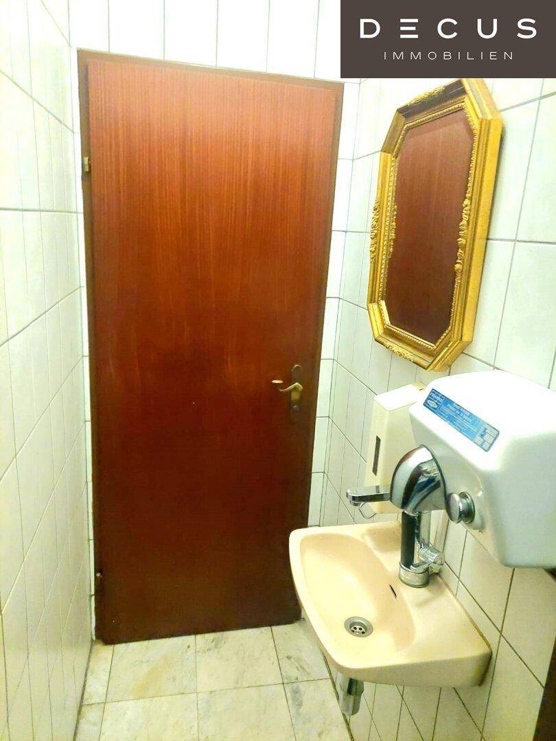 Ein Waschbereich in einem WC mit Waschbecken, Spiegel und Handtrockner, umgeben von weißen Fliesen.