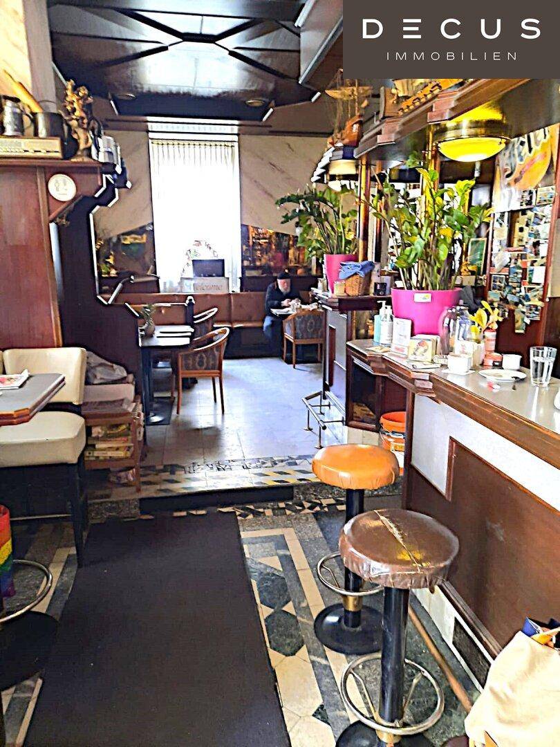 Eine Bar mit Hockern und einem angrenzenden Gastraum mit Tischen und Sitzgelegenheiten, beleuchtet durch Fenster.